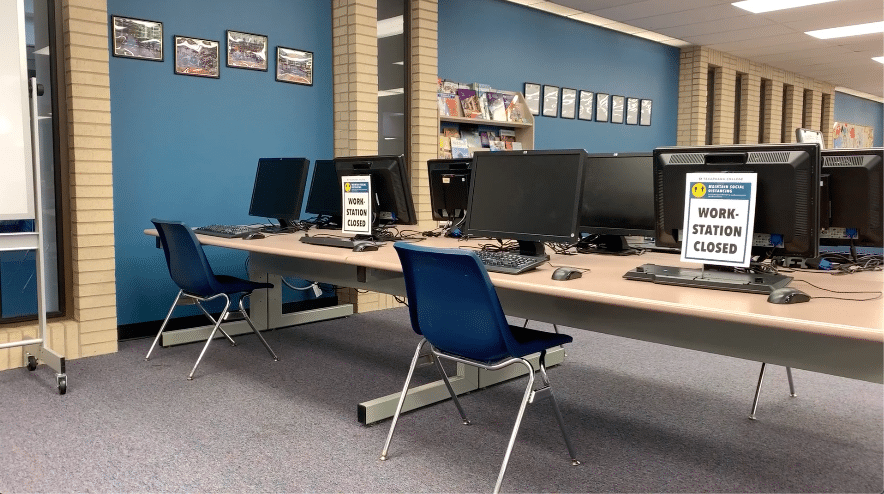 A row of computers in a library or computer lab, with some monitors displaying signs that read Workstation Closed. Blue chairs are placed at each workstation. Shelves with books and pictures line the walls in the background.