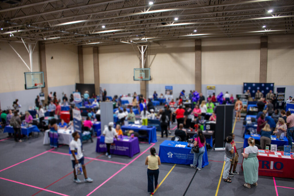 A busy indoor gym hosts a community fair with people visiting various booths, some with colorful tablecloths and banners. Basketball hoops are visible on the walls.