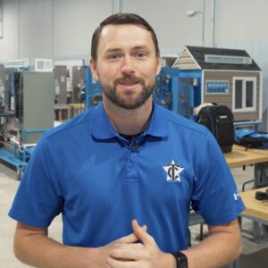 A man with short brown hair and a trimmed beard, wearing a blue polo shirt with a white logo, stands indoors in a workshop or classroom with tables and equipment in the background.