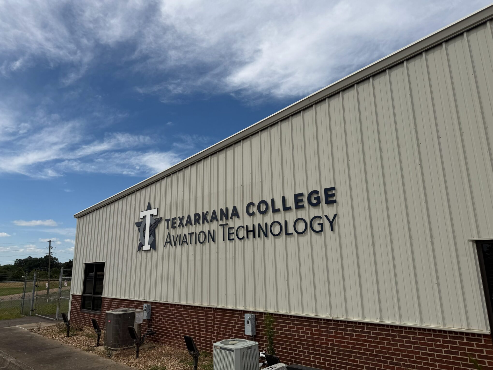 A beige and brick building with a sign that reads Texarkana College Aviation Technology under a partly cloudy blue sky. The building has HVAC units and utility boxes along the side.