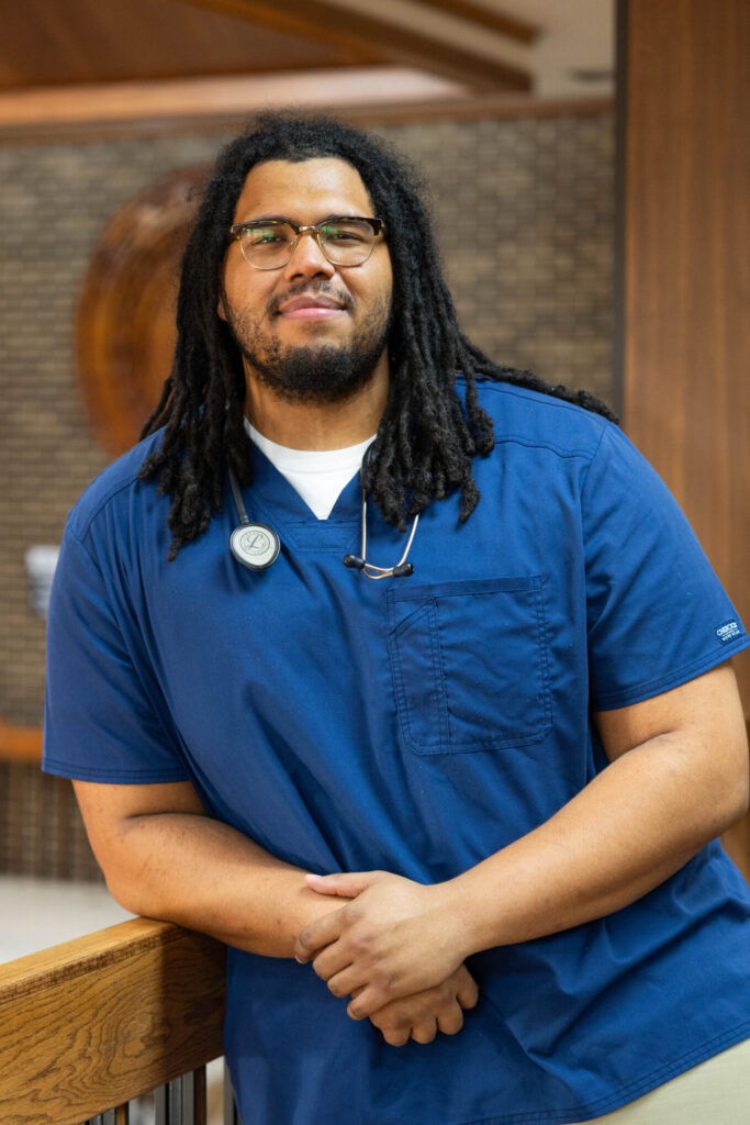 A man wearing blue medical scrubs and glasses stands indoors, leaning on a railing with a stethoscope around his neck. He has long, dark dreadlocks and is smiling slightly. The background features a brown brick wall.