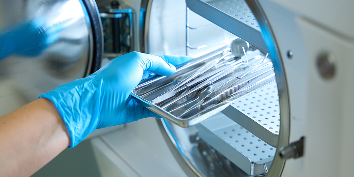 A gloved hand places a metal tray with surgical instruments into a sterilization machine, likely an autoclave, for cleaning and sterilizing the tools.