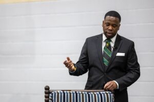 A man in a black suit and striped green tie speaks while standing behind a striped chair, gesturing with his hand. The background is plain and gray.