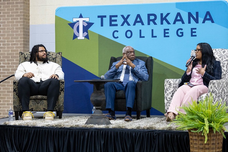 Three people sit on stage having a discussion in front of a blue Texarkana College backdrop. One person is speaking into a microphone while the others listen. A plant sits in front of the stage.