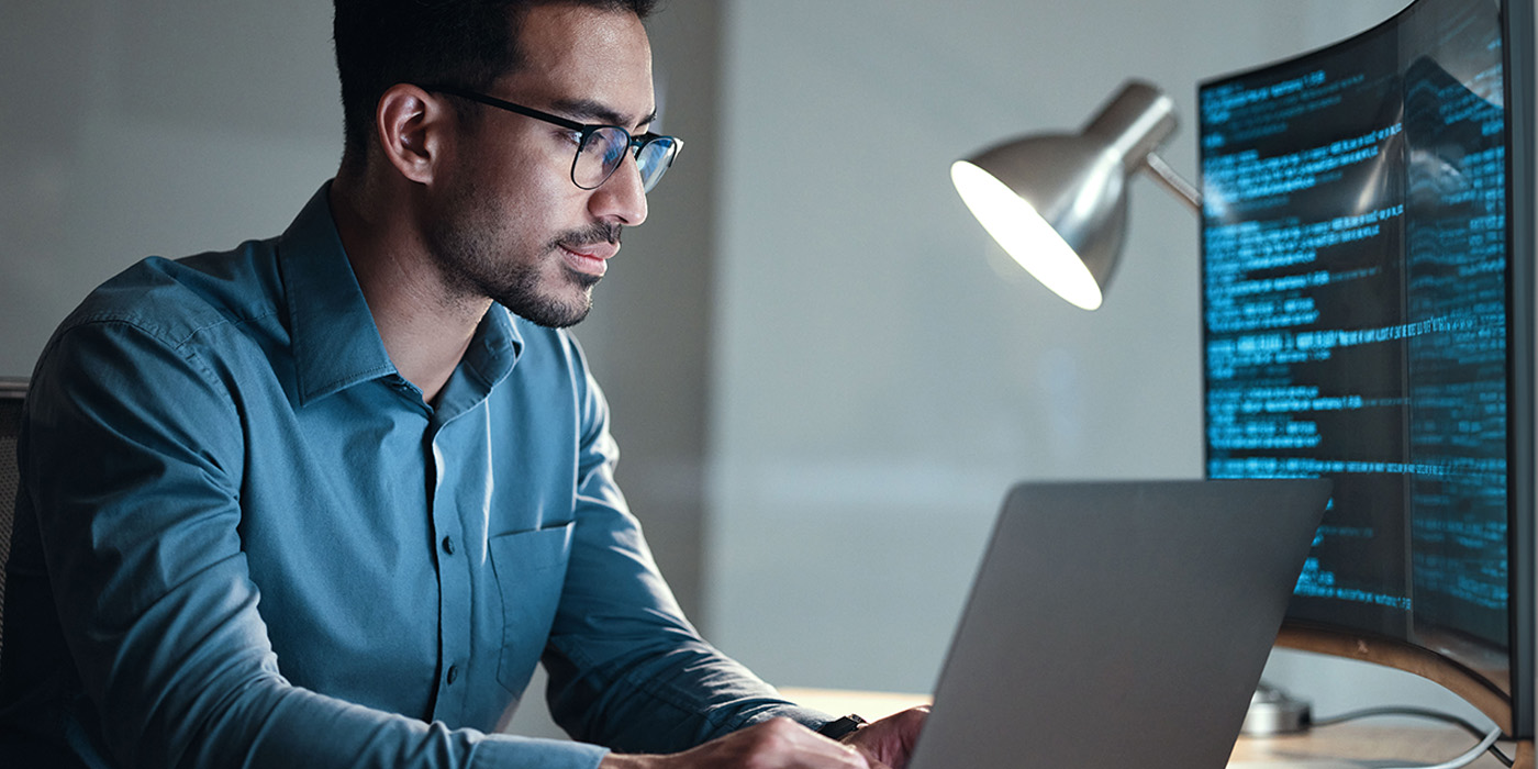 A man wearing glasses and a blue shirt works on a laptop at a desk. He is looking at code displayed on a large monitor, with a desk lamp illuminating the workspace.