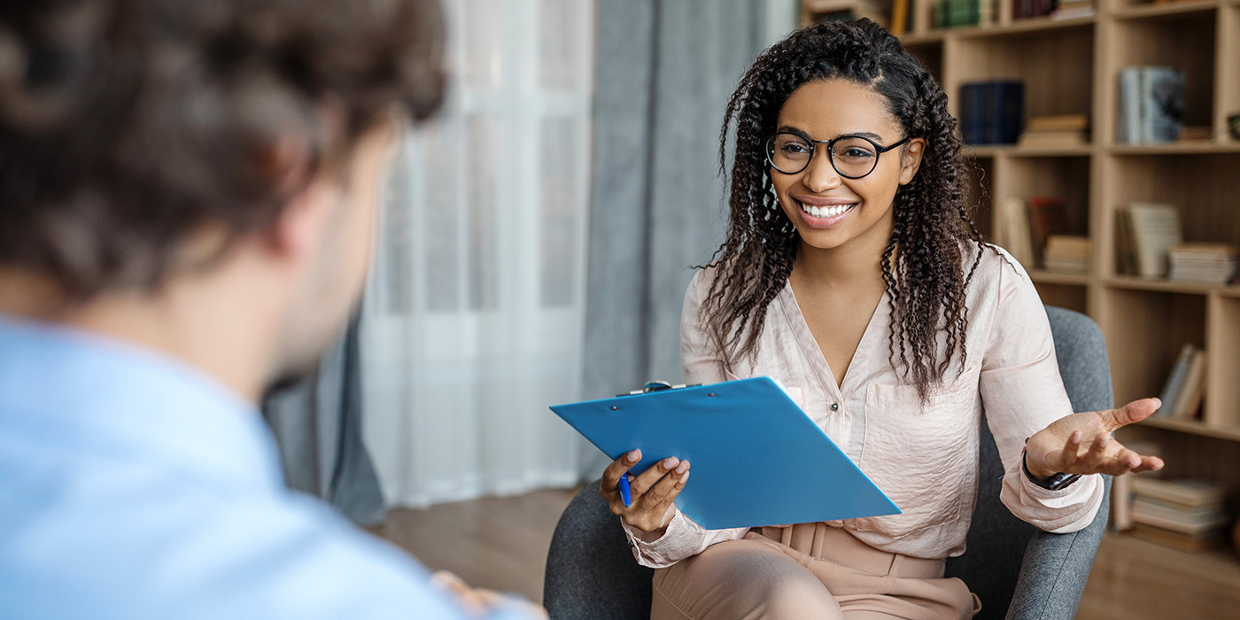 A smiling woman holding a blue clipboard talks with a man in an office setting. She appears engaged and friendly. Bookshelves and a window are visible in the background.