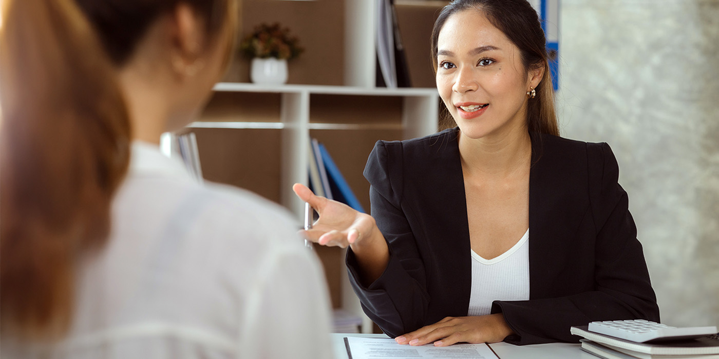 Two women sit across from each other at a desk in an office. One woman, dressed in a black blazer, is smiling and gesturing as she speaks, while the other woman listens with her back to the camera.