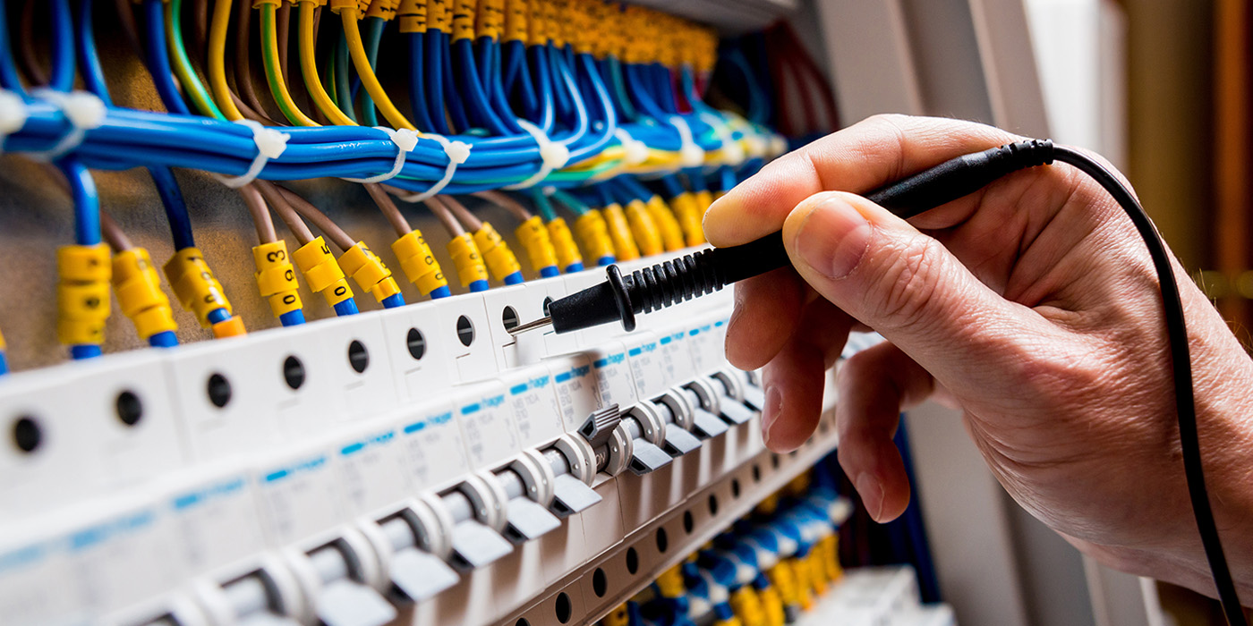 A hand uses a probe to test connections in an electrical panel filled with wires and circuit breakers. The wires are color-coded and organized above the row of white breakers.