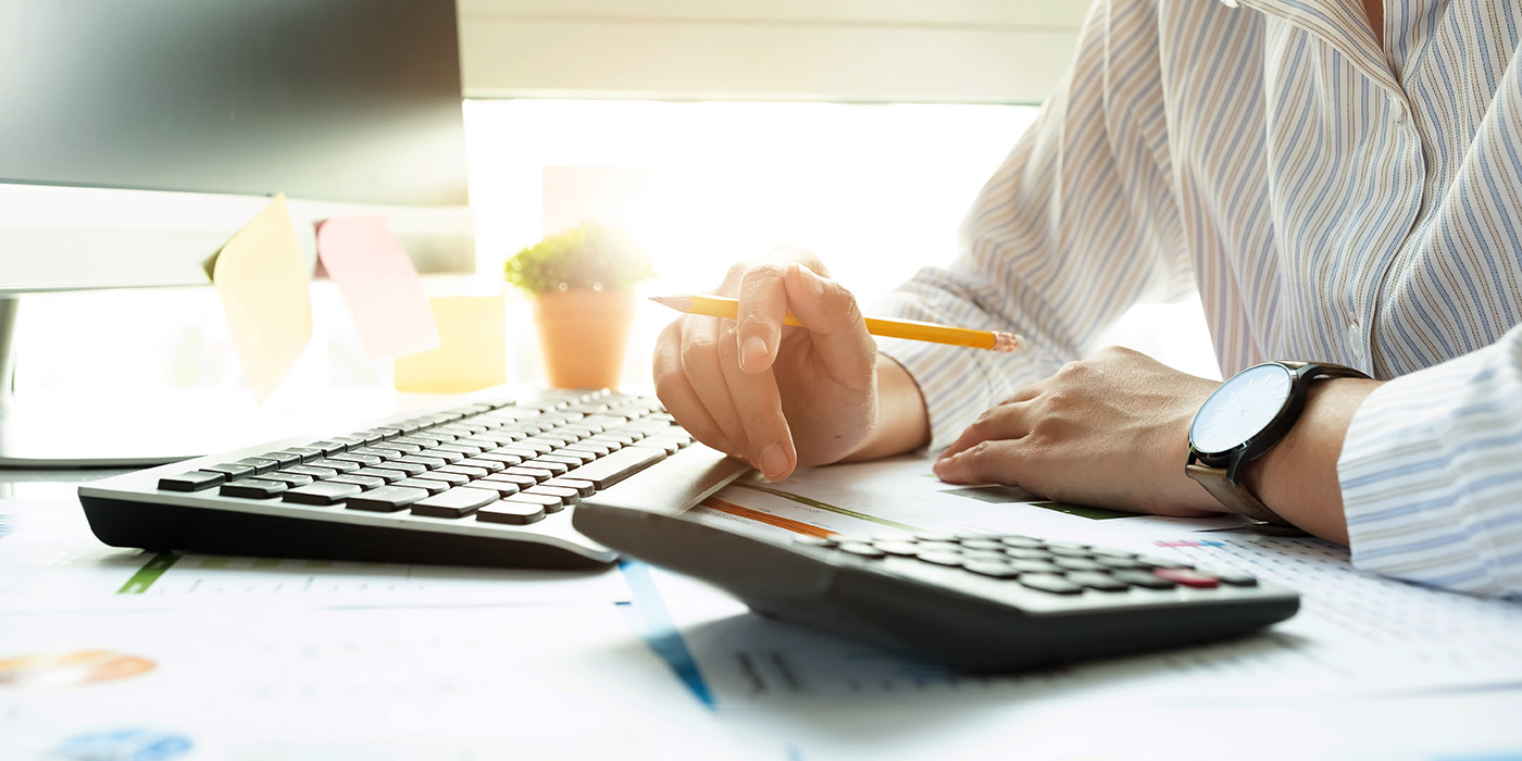 A person in a striped shirt works at a desk with a keyboard, calculator, and documents, holding a pencil. Sunlight comes through a window and there are sticky notes and a small potted plant in the background.