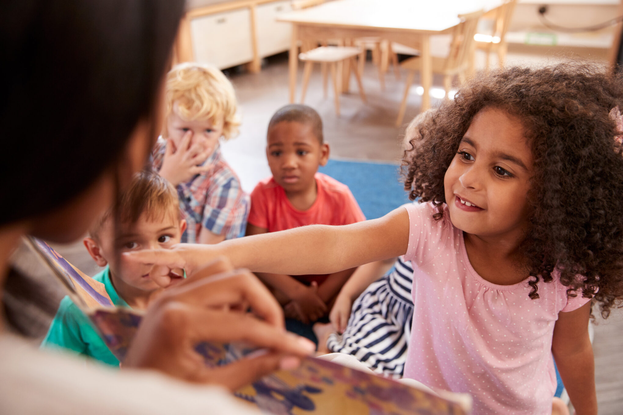 A young girl excitedly points at a picture in a book being read by an adult to a group of attentive children sitting on the floor in a classroom.