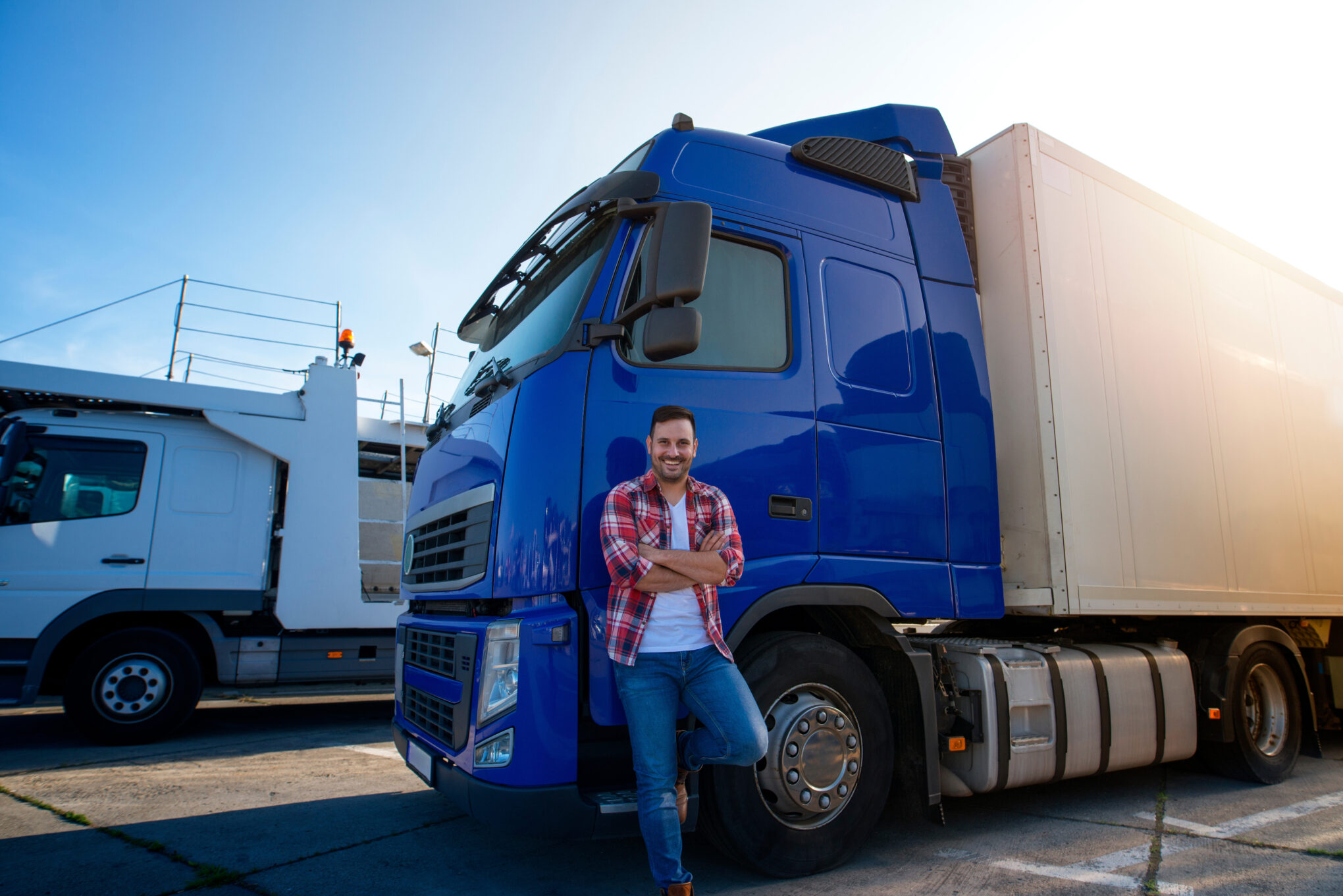 A smiling man in a plaid shirt and jeans stands with arms crossed in front of a blue semi-truck, parked outdoors on a sunny day, with another white truck visible in the background.