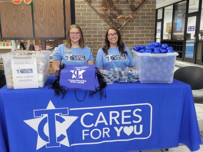 Two women in matching blue TC Cares For You shirts sit at a table with giveaways like bags, buttons, and blue stress balls. The table has a Texarkana College logo and is set up in a public indoor area.