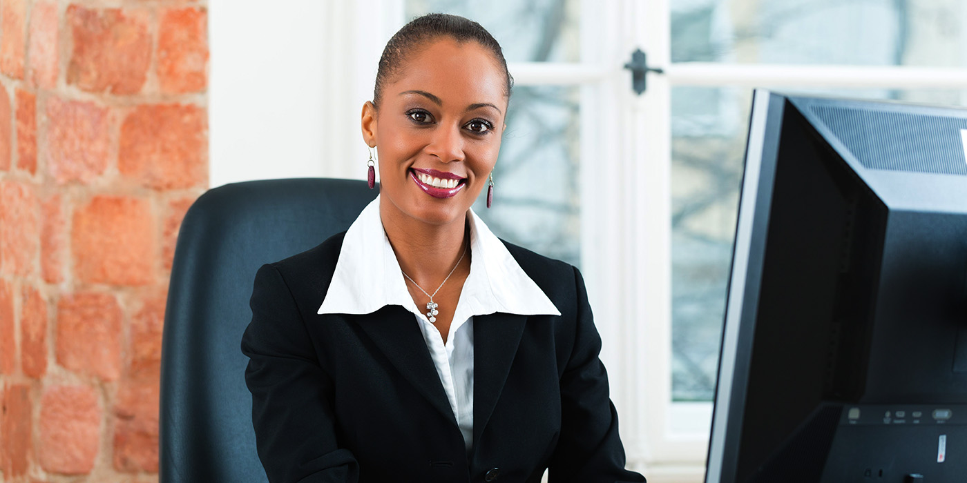 A woman in a black blazer and white blouse sits at a desk in an office, smiling at the camera. She is in front of a computer, with a window and brick wall in the background.