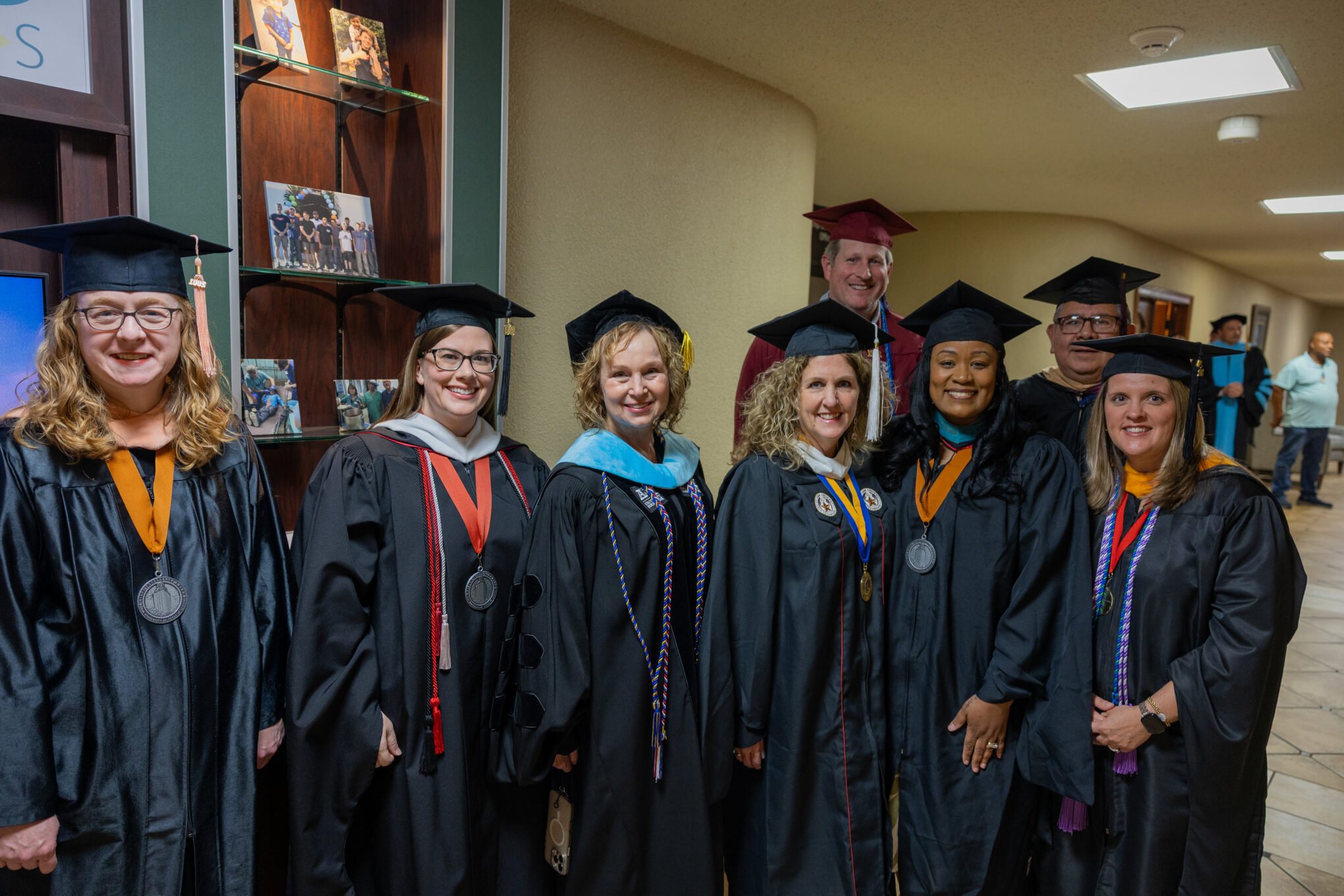 A group of people in graduation caps and gowns stand indoors, smiling for the camera. Some wear medals and academic regalia, and there are photos displayed on shelves in the background.