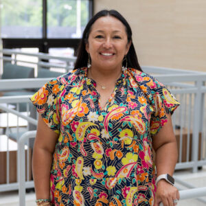 A woman wearing a colorful, patterned dress stands smiling and holding a railing inside a modern, bright building with large windows and light-colored walls.