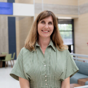 A woman with long brown hair wearing a green and white striped dress stands indoors, smiling at the camera. The setting appears modern with light-colored walls and large windows.