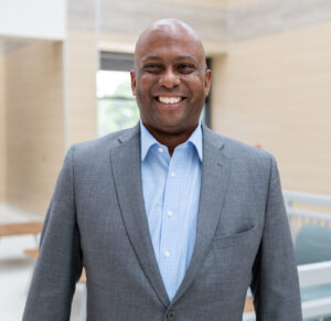 A smiling man in a gray suit and blue shirt stands indoors in a modern, bright building with light-colored walls and furniture in the background.