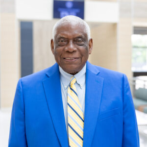 An older man in a bright blue suit jacket, light blue shirt, and yellow striped tie smiles while standing in a modern, sunlit lobby with large windows and seating in the background.