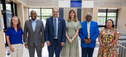 Six professionally dressed people stand in a row, smiling, in a bright indoor hallway with large windows and a sign that reads LAW PRACT.