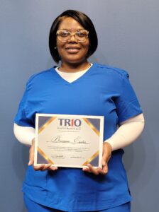 A woman wearing blue medical scrubs and glasses stands against a blue wall, smiling and holding a TRIO Educational Opportunity Center certificate with her name on it.