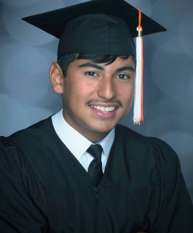 Smiling young man wearing a black graduation cap and gown with a white shirt and black tie, posing in front of a gray backdrop. An orange and white tassel hangs from his cap.