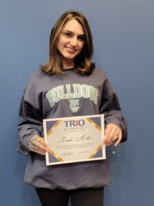 A woman with shoulder-length brown hair, wearing a gray BULLDOGS sweatshirt, stands in front of a blue wall and smiles while holding a TRIO Educational Opportunity Center certificate.