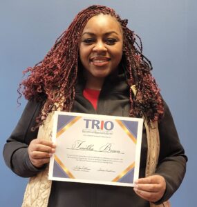 A person with long braids is smiling and holding a certificate from the TRIO Educational Opportunity Center against a plain blue background.