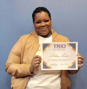 A smiling woman in a tan jacket holds a TRIO Educational Opportunity Center certificate against a blue wall.