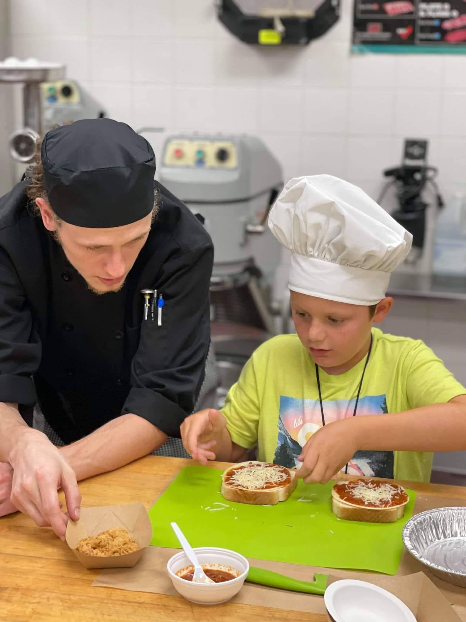 A chef and a young boy in chef hats are making mini pizzas together in a kitchen. The chef guides the boy, who is sprinkling cheese on pizza sauce, while bowls of ingredients sit on the table.