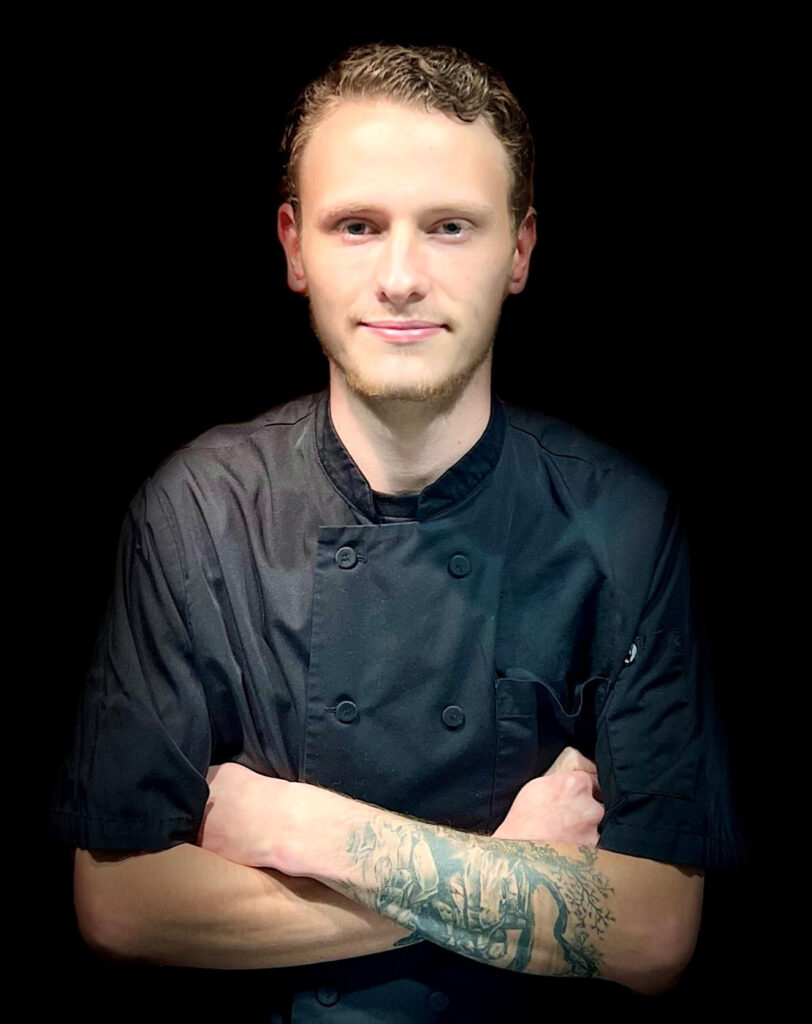 A man in a black chefs jacket stands against a black background with his arms crossed, showing a tattoo on his forearm. He has short, wavy hair and a slight smile.