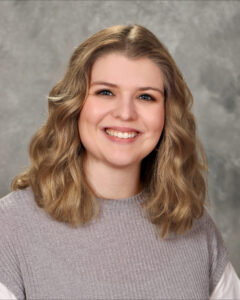 A woman with shoulder-length wavy blonde hair smiles at the camera. She is wearing a light gray ribbed top and is posed in front of a neutral gray, textured background.