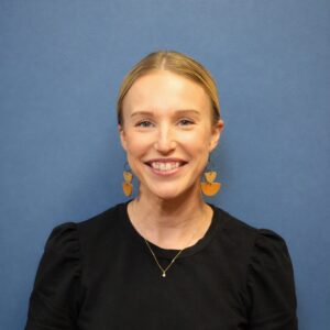 A woman with blonde hair in a low bun, wearing a black top, statement earrings, and a simple necklace, smiles in front of a plain blue background.