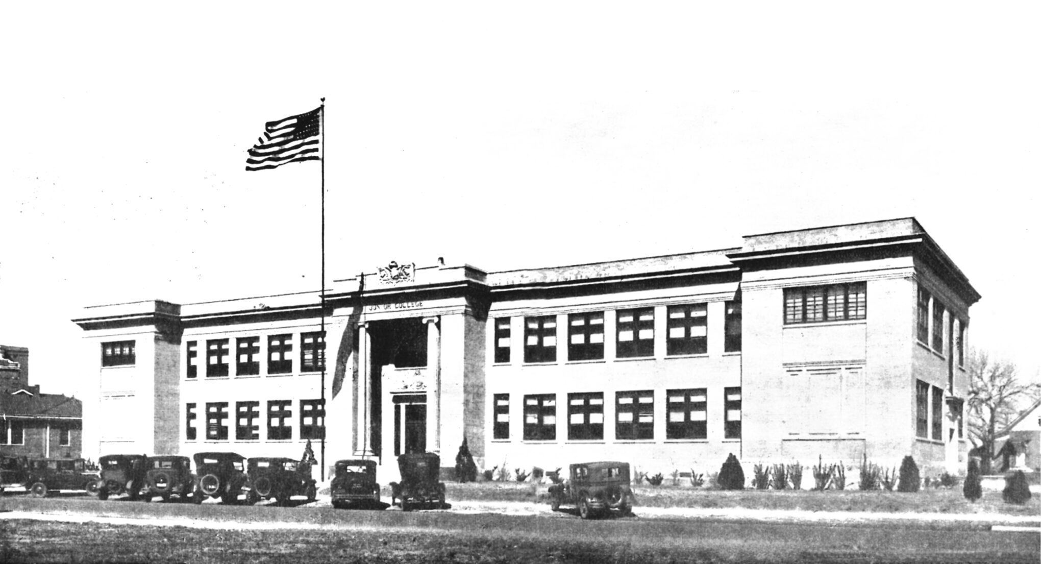 Black-and-white photo of a two-story school building with an American flag on a pole in front. Vintage cars are parked along the street, and the main entrance features columns.
