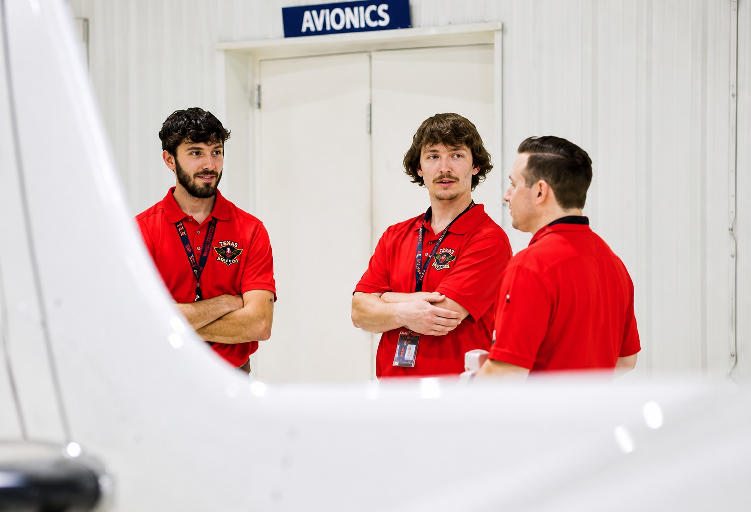 Three men in red shirts stand and talk in front of a door labeled Avionics inside a white room, with part of an aircraft visible in the foreground.