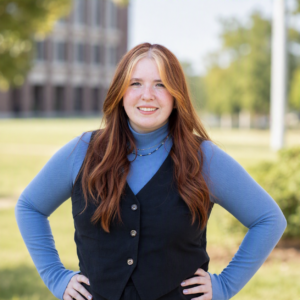 A young woman with long red hair, wearing a blue turtleneck and black vest, stands outdoors with her hands on her hips, smiling at the camera. The background shows greenery and a blurred building.