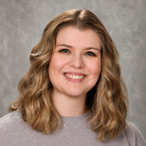A woman with wavy, shoulder-length blonde hair smiles at the camera, wearing a light gray sweater. The background is a neutral gray.