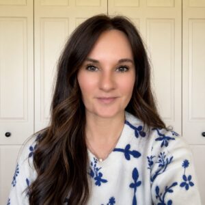 A woman with long brown hair wearing a white sweater with blue floral patterns smiles at the camera. She is standing indoors in front of cream-colored paneled doors.