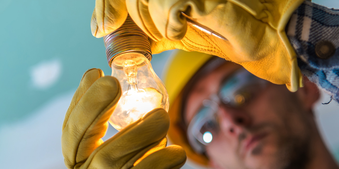 A person wearing yellow safety gloves and a hard hat installs a glowing light bulb, focusing on the hands and the illuminated bulb.