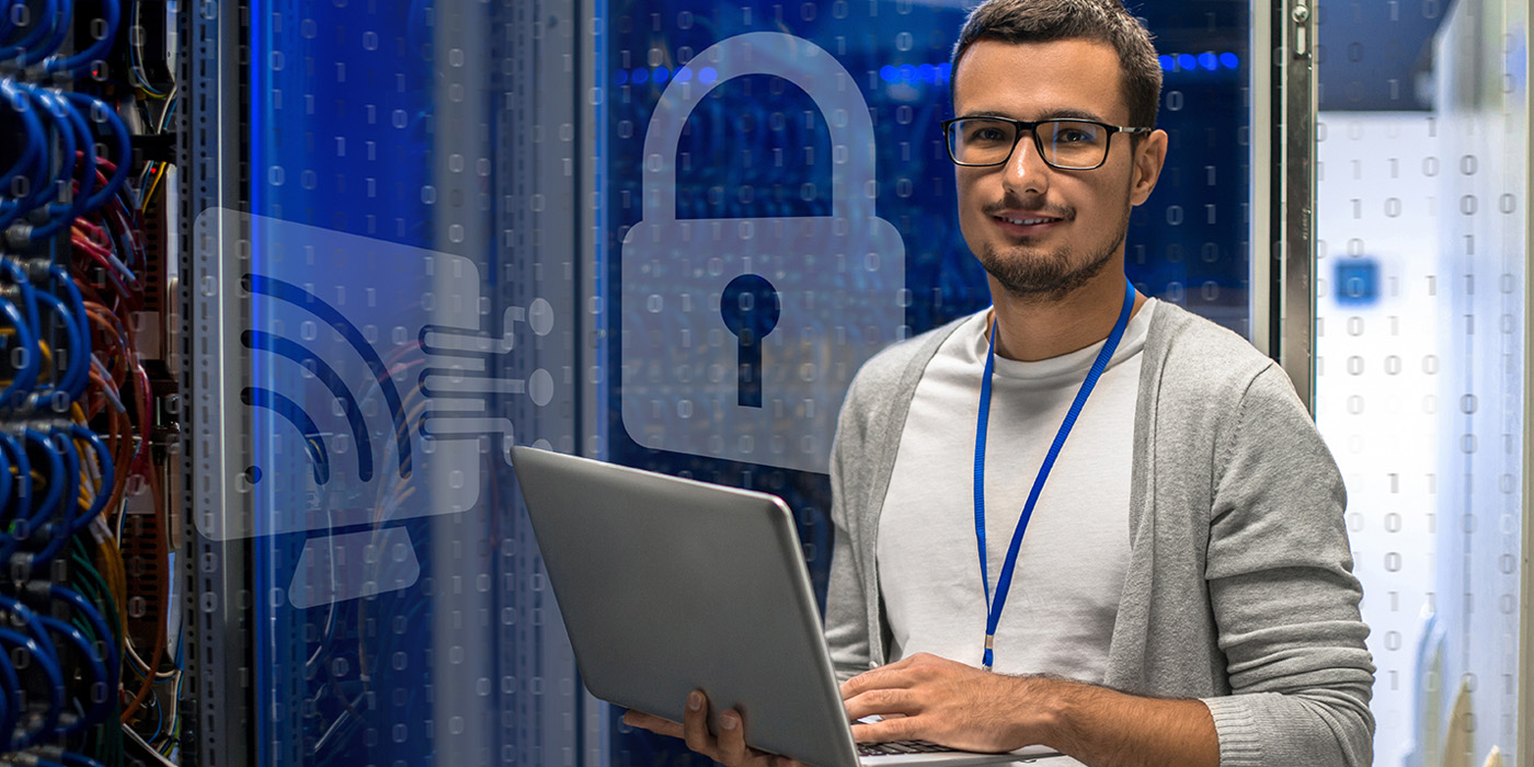 A man with glasses holds a laptop, standing in a server room. Digital icons of a lock and Wi-Fi signal are overlaid, symbolizing cybersecurity and network protection. He is wearing an ID badge and smiling.