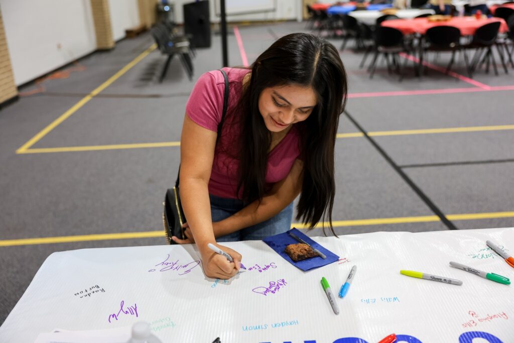 A woman with long dark hair signs a large white banner with colorful markers. She is smiling and wearing a pink shirt. Tables and chairs are in the background, and a snack sits on the banner.