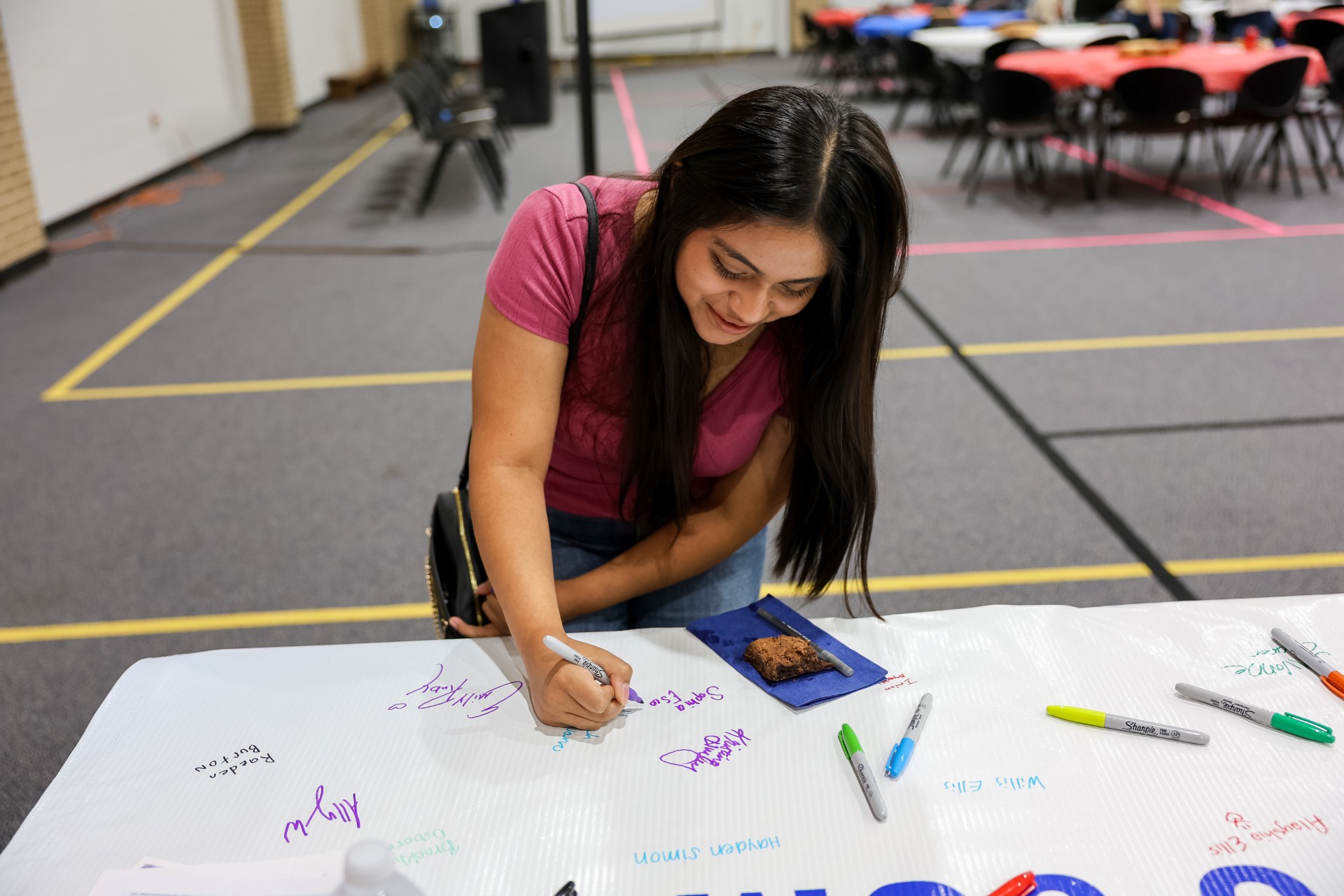 A woman with long dark hair signs a large white banner with colorful markers. She is smiling and wearing a pink shirt. Tables and chairs are in the background, and a snack sits on the banner.