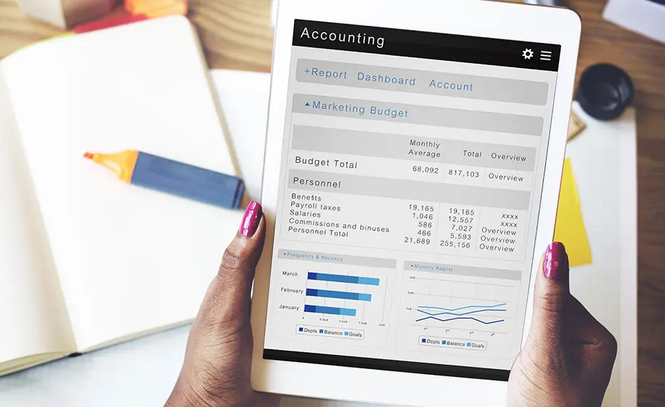 Person holding a tablet displaying an accounting dashboard with budget, personnel expenses, and line graphs. A notebook and highlighter are visible on the desk in the background.
