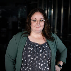 Woman with glasses and long brown hair wears a green blazer over a patterned dress, standing with hands in pockets, smiling at the camera against a dark, blurred background.