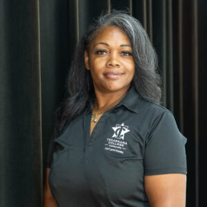 A woman with long dark hair wearing a black polo shirt with a logo stands in front of dark curtains, smiling slightly. She has tattoos on her forearms and her hands are clasped in front of her.