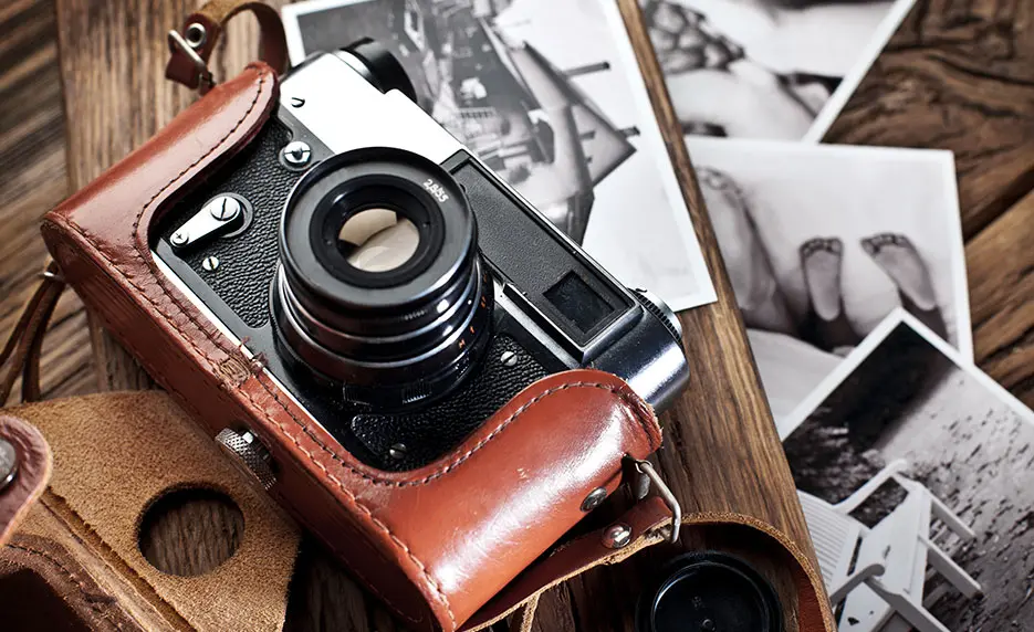 A vintage camera in a brown leather case sits on a table surrounded by black-and-white photos. The lens faces forward, and the case is partially open, revealing part of the camera body and nearby photo prints.