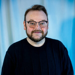 A man with glasses, a beard, and short hair is wearing a black shirt and standing in front of a light blue background, smiling softly at the camera.