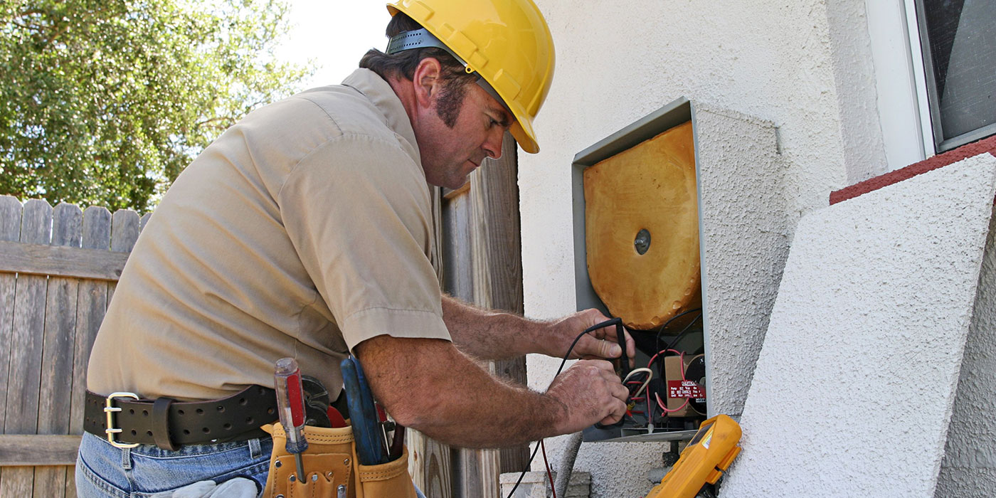 An electrician wearing a yellow hard hat and tool belt repairs wiring in an outdoor electrical panel attached to a house.