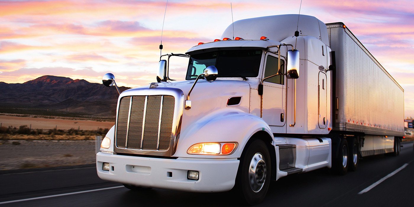 A white semi-truck with a trailer drives on a highway at sunset, with mountains and colorful sky in the background.
