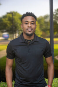 A young man wearing a black polo shirt and a gold cross necklace standing outdoors.
