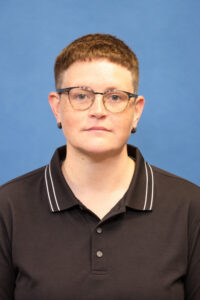 Headshot of a person with short brown hair and glasses, wearing a black polo shirt against a blue background.